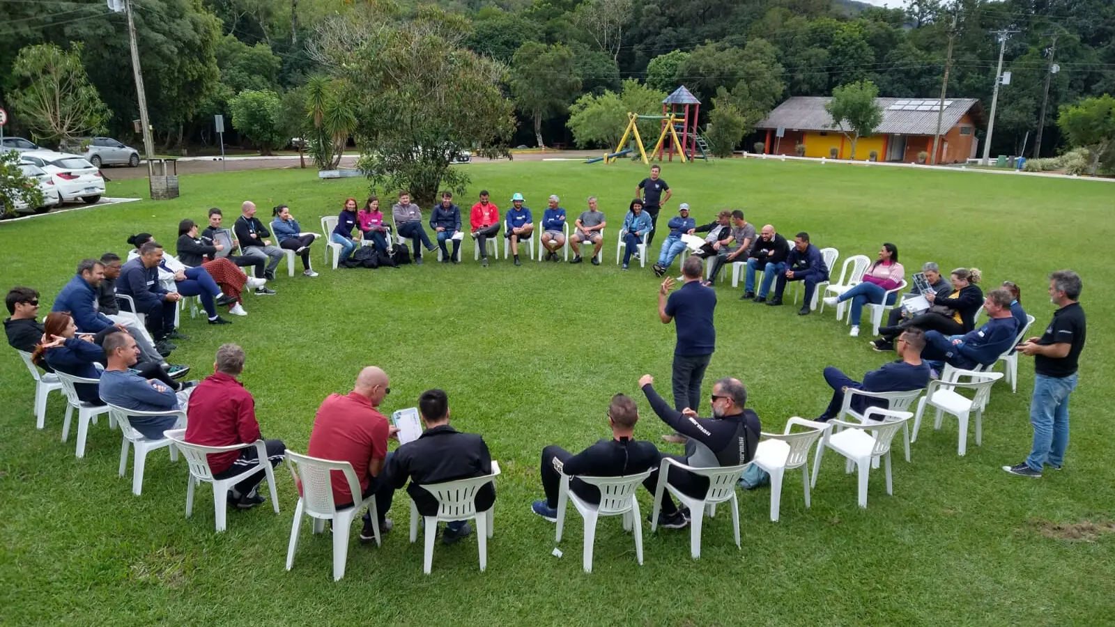 Encontro em círculo de participantes em uma reunião ao ar livre, rodeados por natureza e árvores, com cadeiras brancas dispostas de forma organizada.