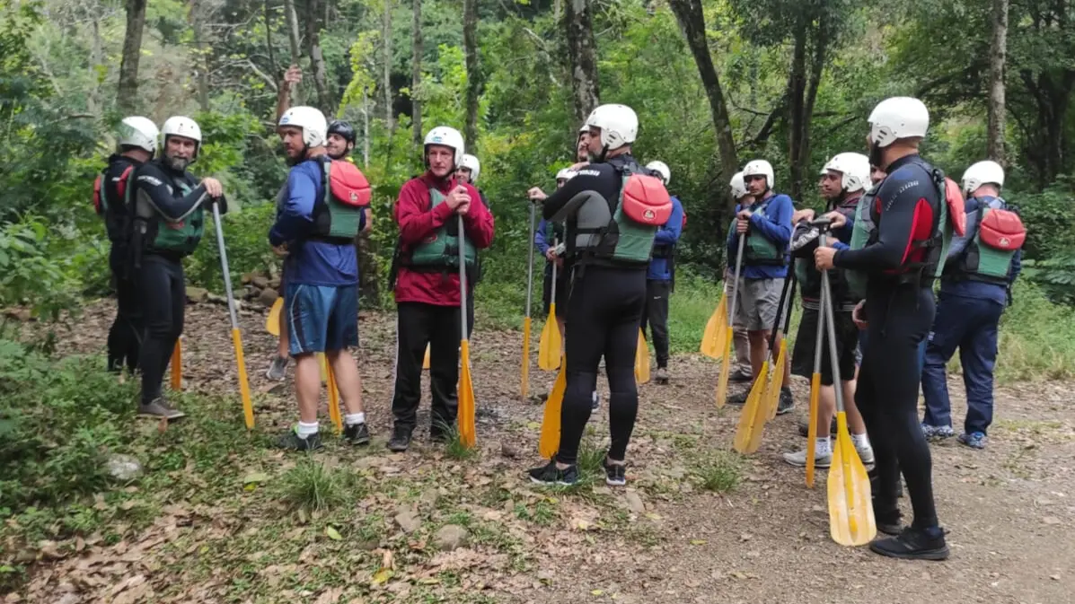 Grupo de pessoas reunidas em um ambiente natural, se preparando para uma atividade de rafting. Todos usam coletes e capacetes de segurança, segurando remos.
