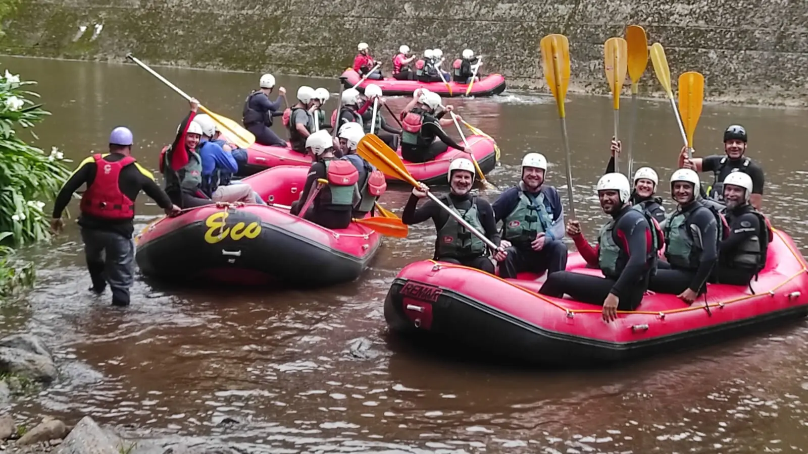 Grupo de pessoas se divertindo em um passeio de rafting em embarcações infláveis em um rio. Eles estão equipados com coletes salva-vidas e remos, prontos para a aventura.