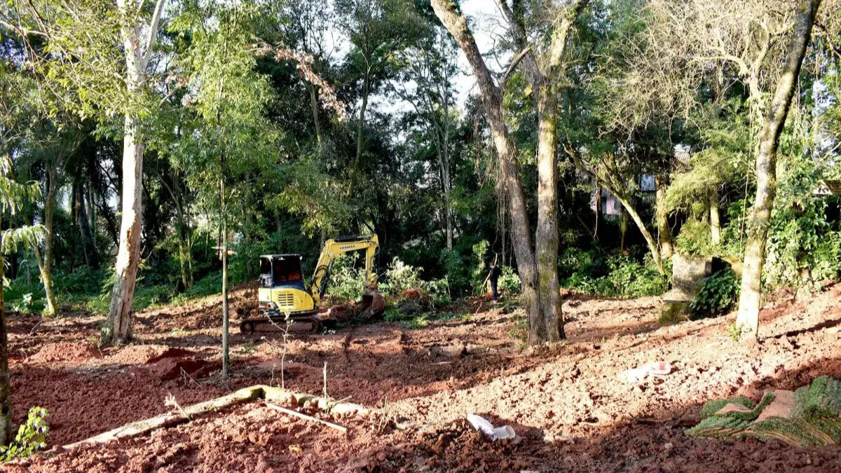 Imagem de uma área com máquinas de construção trabalhando em um terreno repleto de vegetação e árvores. O local mostra sinais de preparação para construção.