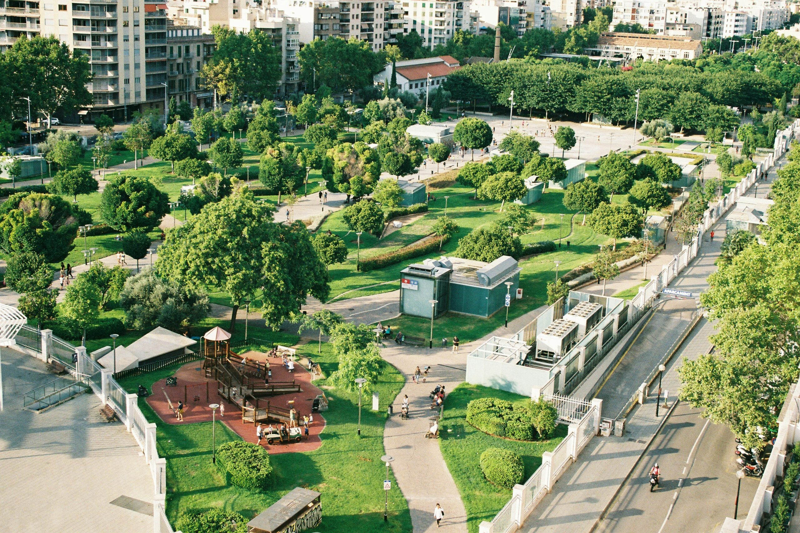 Vista aérea de um parque urbano com áreas verdes, playgrouds e caminhos para pedestres em uma cidade moderna.